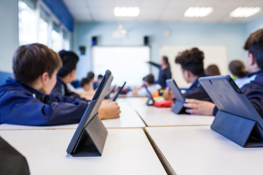 A group of children seated at tables faces the teacher in the front of the room with tablets in front of them.