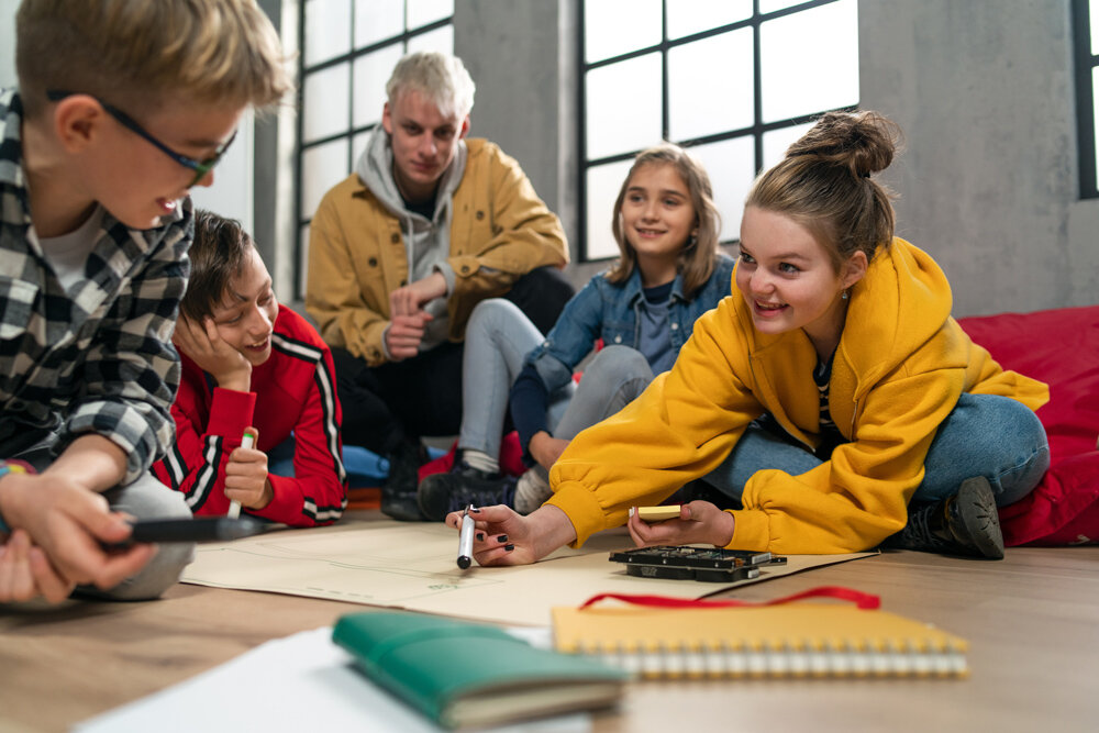 A group of students sit together writing on a big poster board with notebooks scattered around on the ground.