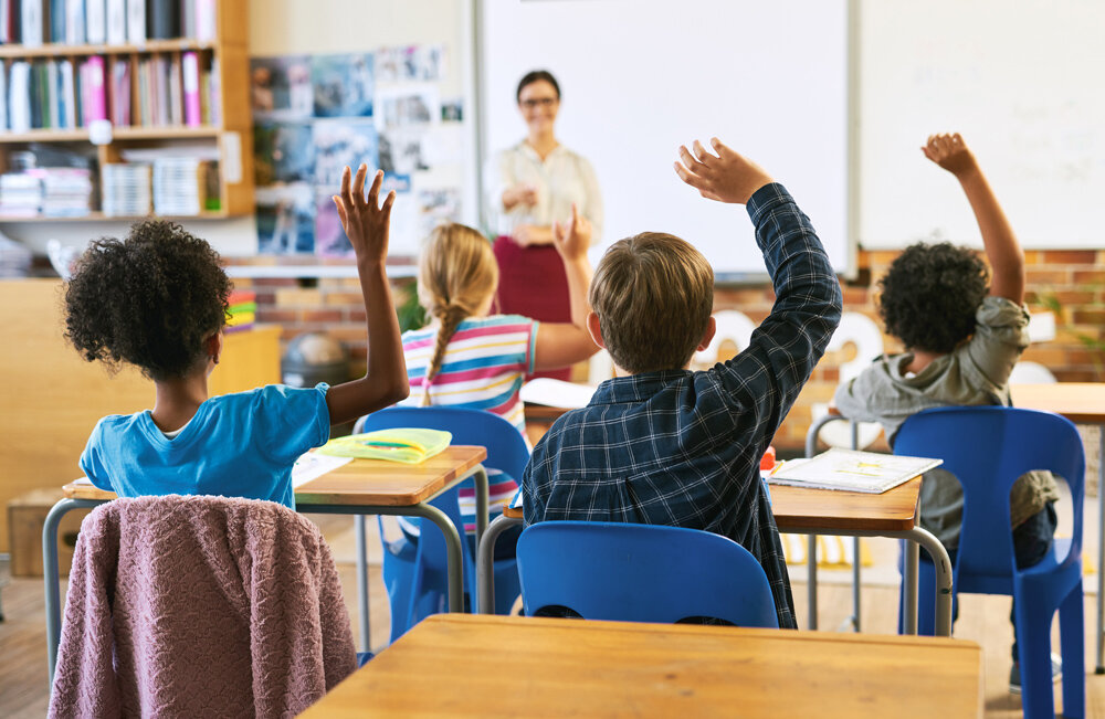 A group of students sits in a classroom raising their hands while the teacher stands in front of them.